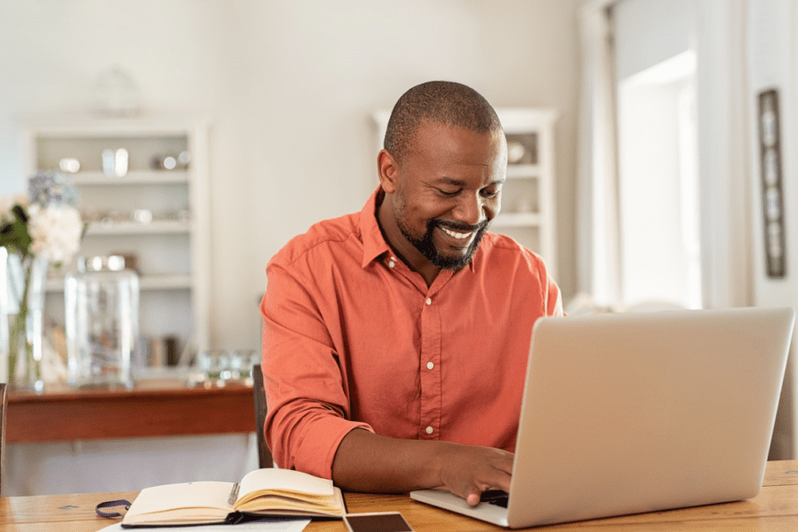 Smiling man working on laptop from home wearing red shirt with kitchen in background