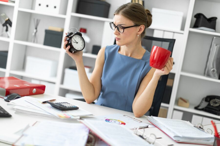 A young girl is sitting at a table in her office, holding an alarm clock and a red cup.