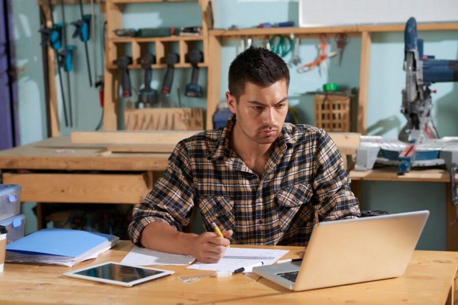 Photograph of a person sitting at a wooden workbench in a workshop, using a laptop and surrounded by papers.