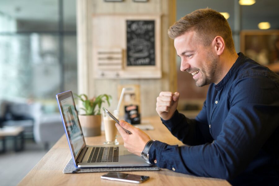 Photograph of a man sitting at a wooden counter in a cafe, using a smartphone while a laptop is open in front of him.